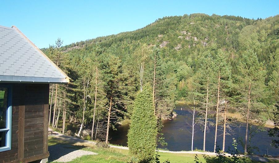 Brun laksehytte, grønn hage og vann nedenfor. Skog bak vannet, blå himmel.
