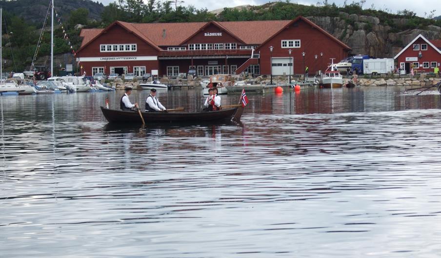Liten trebåt på vannet med 2 menn og 2 kvinner i bunader, og det norske flagget på tuppen av båten foran den røde Lindesnes Hytteservice bygningen.