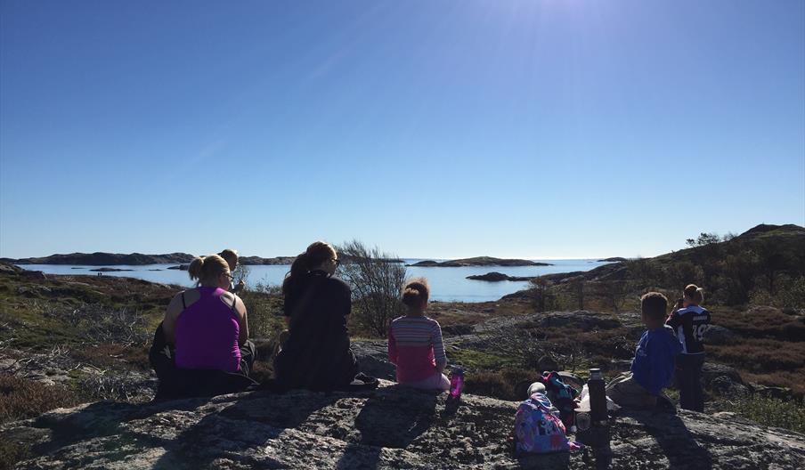 Bilde av seks mennesker som sitter på stein og ser utover mot havet, sola skinner ned på dem, en dame tar bilde av utsikten