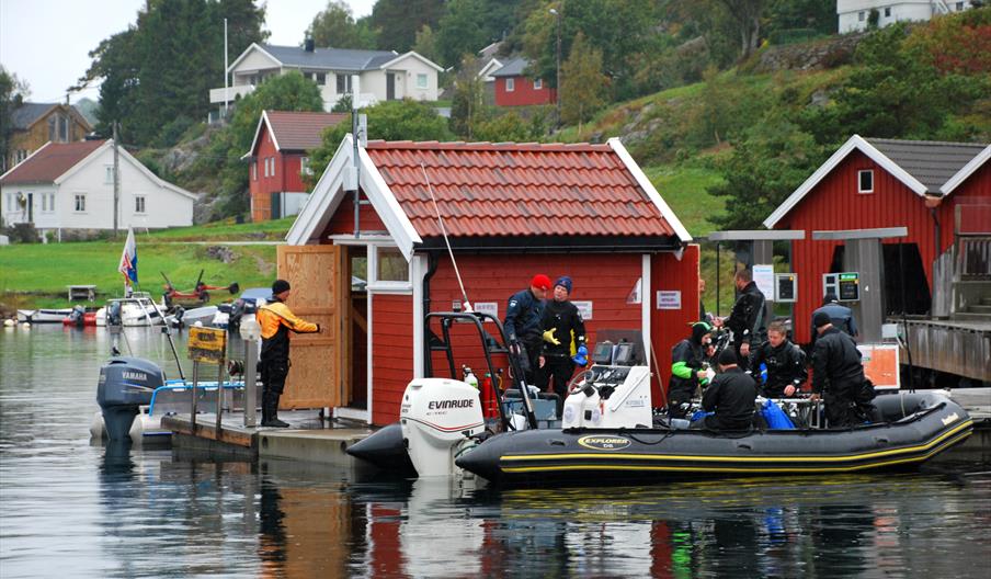Bilde av en rød bod på en brygge med minst 8 dykkere som gjør seg klare for å komme seg på en svart båt på Tregde