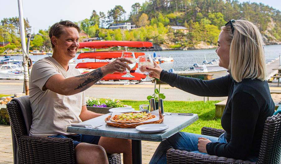 To personer med hvert sitt glass og skåler. Sitter ute i finværet, med en pizza på bordet