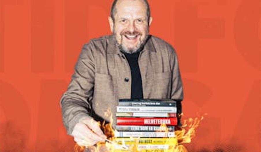 Komiker Freddy Kjensmo smilende med en stabel bøker som står i flammer foran seg.