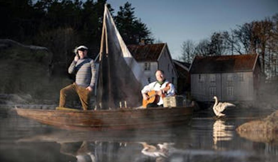 To personer på en liten båt med seil, omgitt av tåke, hvor én spiller gitar og en annen står i karakter som sjømann.