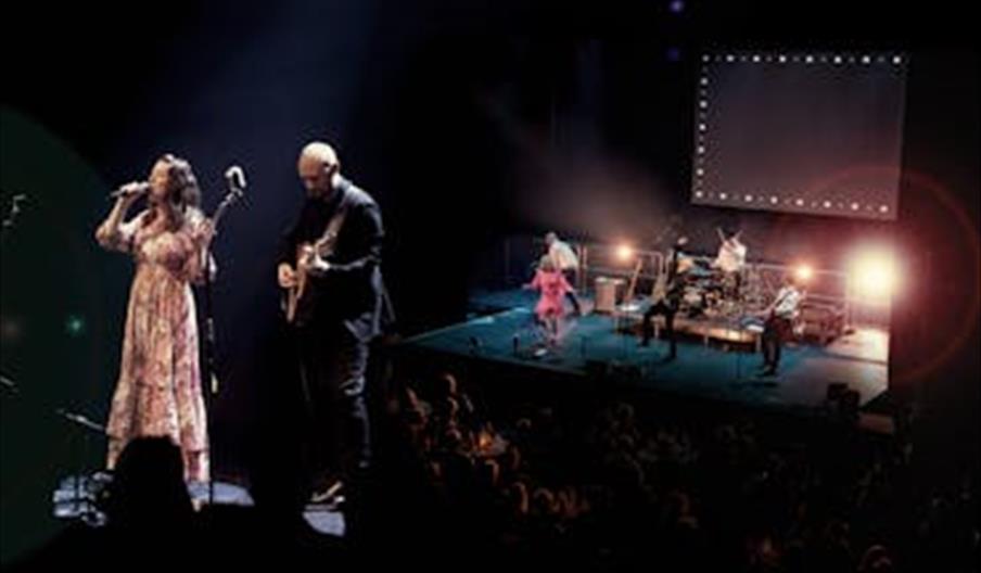 Et band opptrer på en scene med sangere og musikere i varmt scenerlys foran publikum.