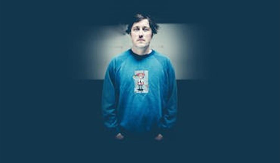 En mann står i en blå genser foran en mørk bakgrunn, brukt i promotering av en konsert.