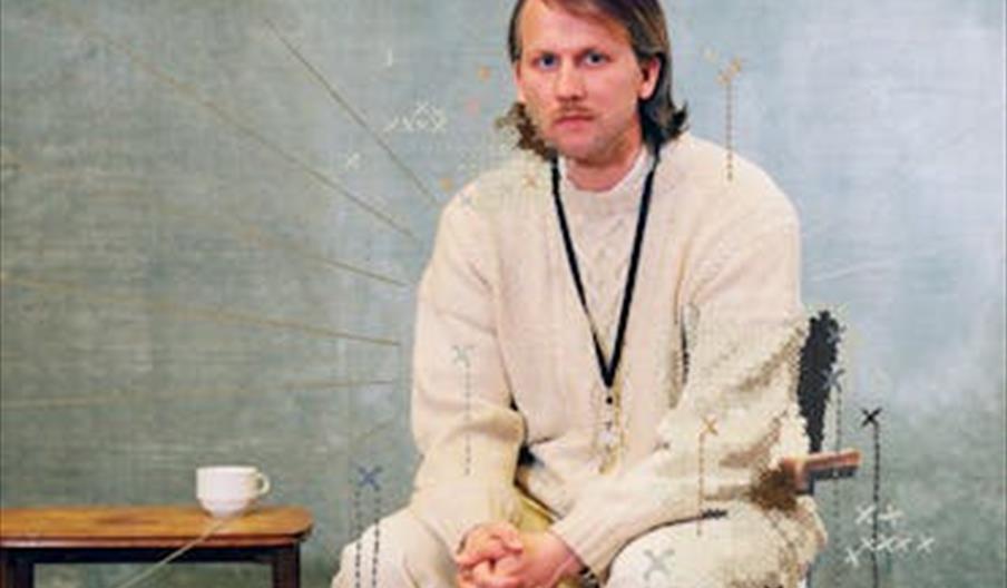 En person sitter på en stol foran en dus grønn bakgrunn med en kopp på et bord ved siden av, brukt i teaterpromotering.