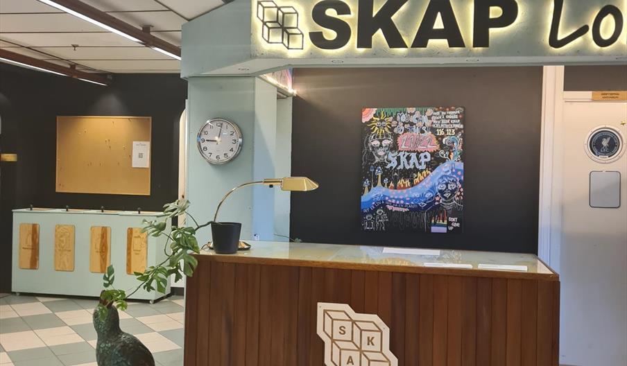 Resepsjonsdisk hos SKAP. Brun disk med SKAP logo på  Blågrå og hvite rutete gulv, blå og svart vegg med svære bokstaver på veggen "SKAP"