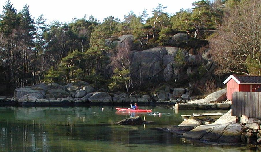 Bilde av en person i rød kajakk i vannet ved en brygge med en rød bygning