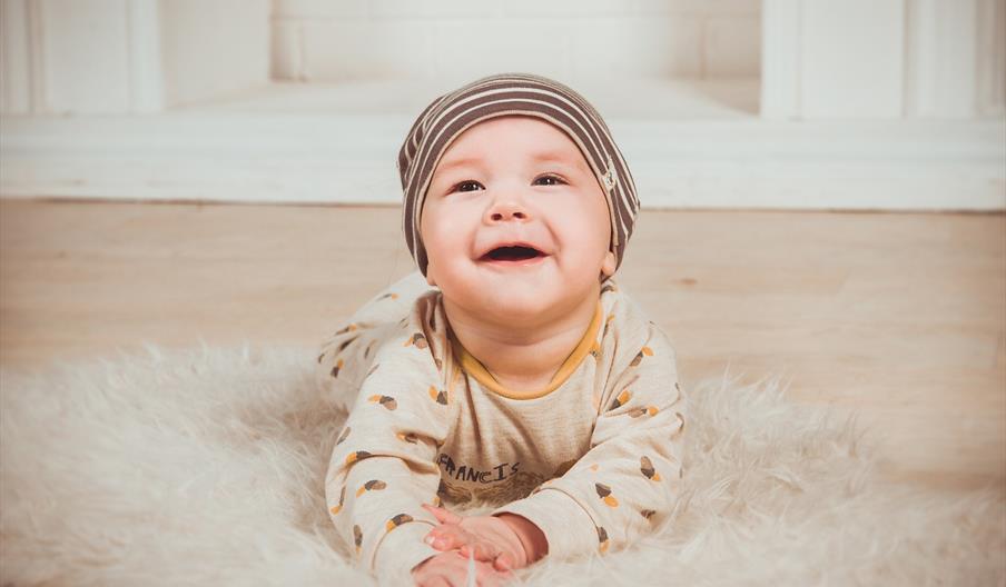 Bilde av en baby liggende på magen, på et teppe på gulvet. Babyen kikker oppover og smiler. Foto: Vika_Glitter