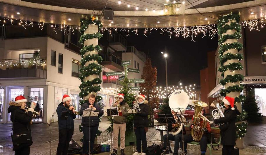 Et korps med barn og unge i nisseluer spiller julemusikk under julebyen Lyngdal i stemningsfull kveldsbelysning.