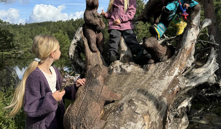 Tre barn som klatrer på en trestokke med utskjæringer av 3 bjørnunger og koser seg på Gluba