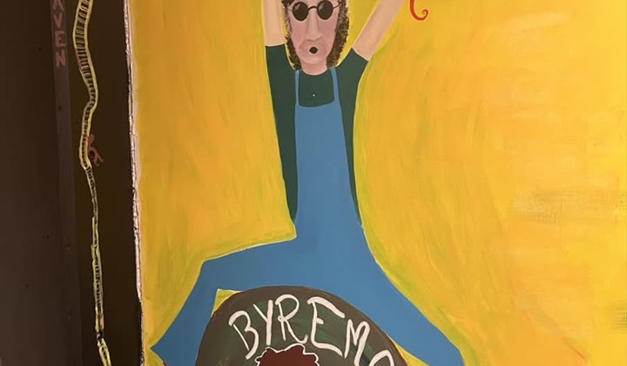 Veggmalt kunstverk med logoen «Byremo Scene» og en figur med solbriller og oppstrakte armer på gul bakgrunn.