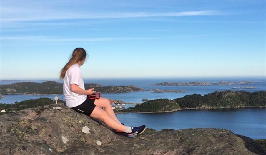 Bilde av en kvinne med hestehale som sitter på fjellet og ser ut mot havet, på havet er det flere fjell