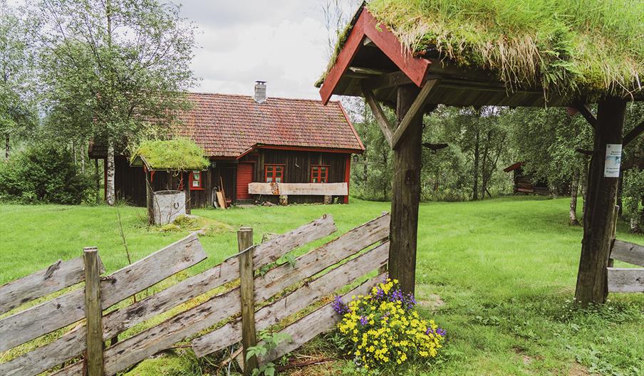 Hagebue i tre med torvtak på, stor gressflekk ed en brun hytte med røde detaljer på listene og døra.