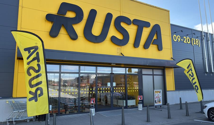 Inngangen til Rusta Lyngdal i Handelsparken, med gul fasade og flagg med logo.