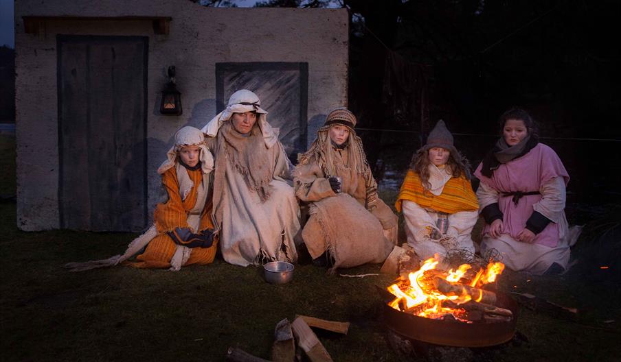 Skuespillere utkledd som gjetere sitter samlet rundt et bål foran en enkel stallvegg under forestillingen «Veien til Betlehem» i Lyngdal.