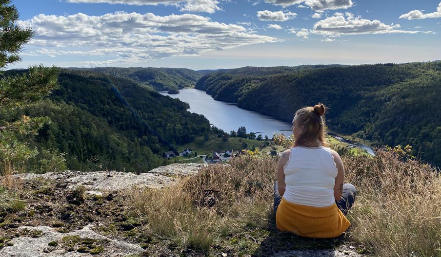 Nomeheia i Øyslebø. En i hvit topp og gul bukse sittende og ser framover. skog til høyre og venstre med elv midt i.