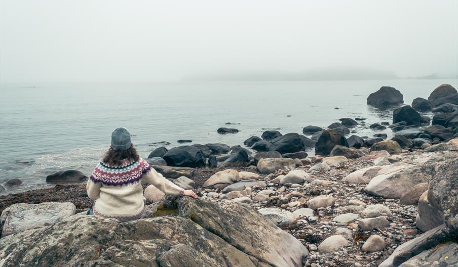 Dame med ullgenser og lue, som sitter på steinene og ser utover vannet