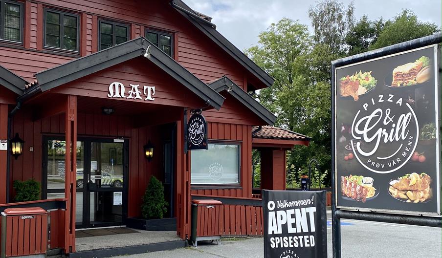 Delvis fasade av inngangen. Rød bygning med hvit skrift "mat" over inngangen. To store plakater med reklame av Mones Pizza & Grill.