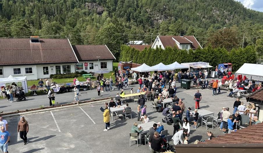 Utsikt over et utendørs marked under Konsmodagen. Mange mennesker samles rundt bord og boder i et gårdsrom, omgitt av telt og små bygg. Stemningen er livlig og omgitt av grønne åser og blå himmel i bakgrunnen.