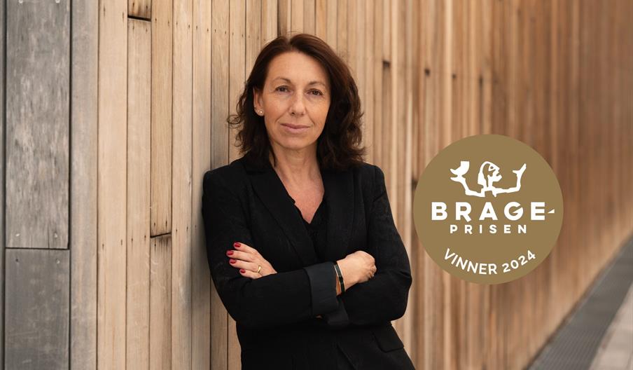 forfatter Kristina Quintano kledd i svart med armene i kors lener seg mot en trevegg. På bilde er også logo av Brageprisen vnner 2024.