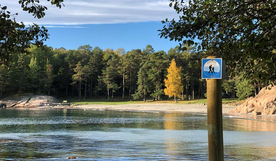 Bukt med strand og trær i bakgrunnen. Et liste skilt med "kyststien" fremme i bildet.