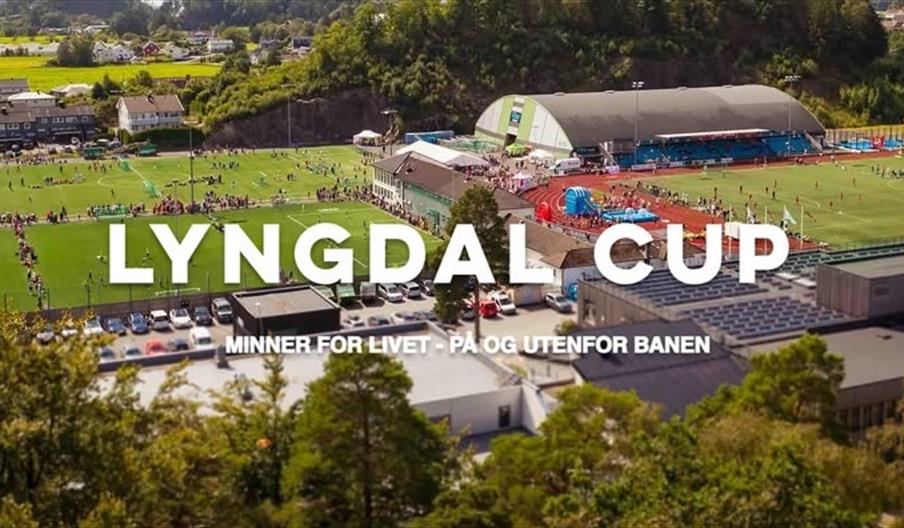 Oversiktsbilde av Lyngdal Stadion fylt med fotballbaner og mennesker under Lyngdal Cup. Midt i bildet står teksten "Lyngdal Cup – Minner for livet, på og utenfor banen". I bakgrunnen sees hall, tribuner og grønne omgivelser.