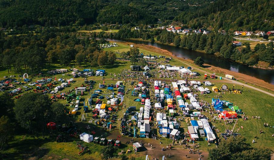 Oversiktsbilde av Dyrskuet i Lyngdal med boder, dyr, maskiner og publikum samlet på et stort uteområde ved elva.