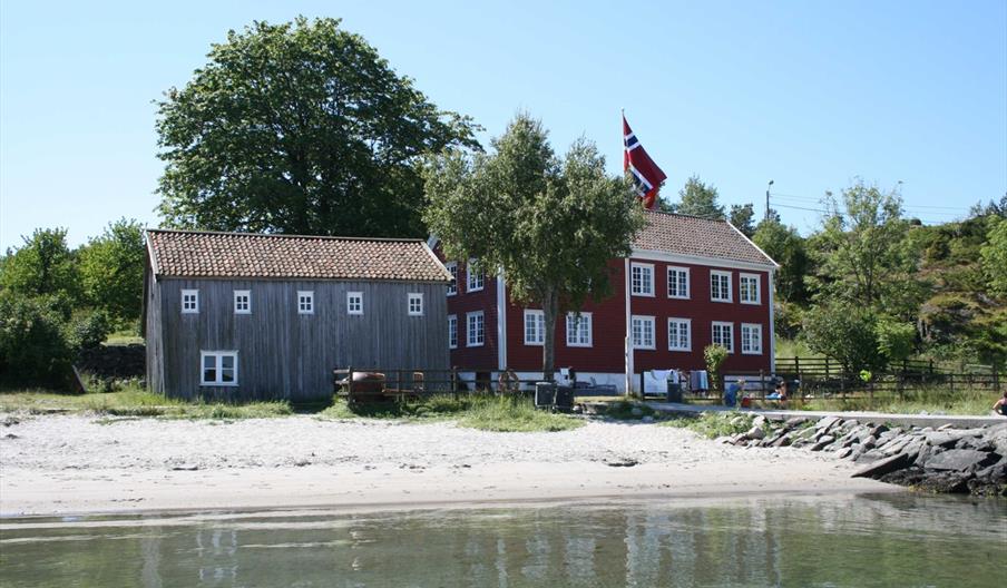 bilde av Merdøgaard og en grå låve. foran bygningene ligger en fin sandstrand.