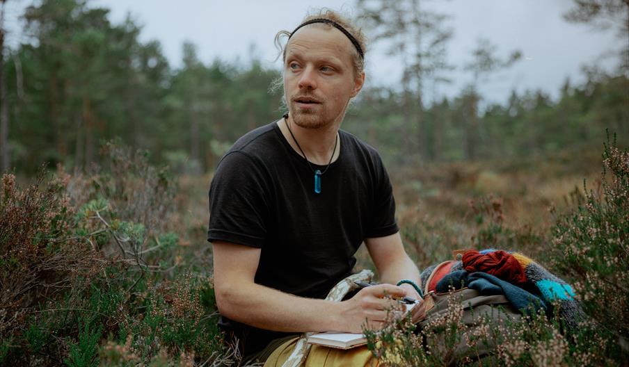 artisten Moddi sittende i lyngen med penn og papir.