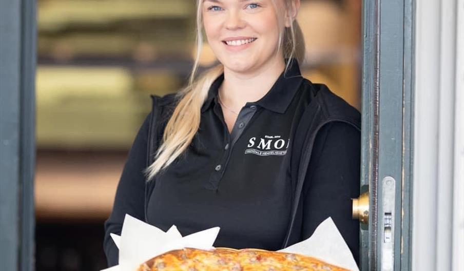 Blid servitør fra SMOI som står i døra og holder en deilig pizza