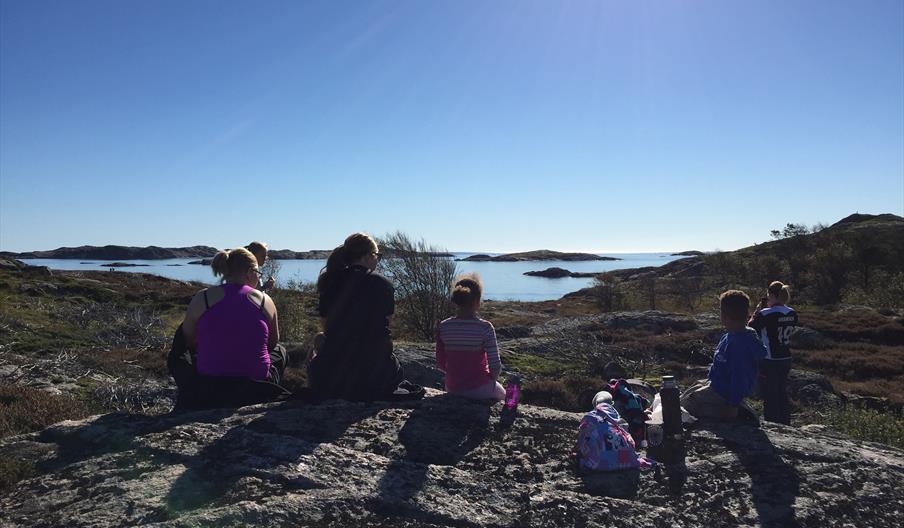 Bilde av seks mennesker som sitter på stein og ser utover mot havet, sola skinner ned på dem, en dame tar bilde av utsikten