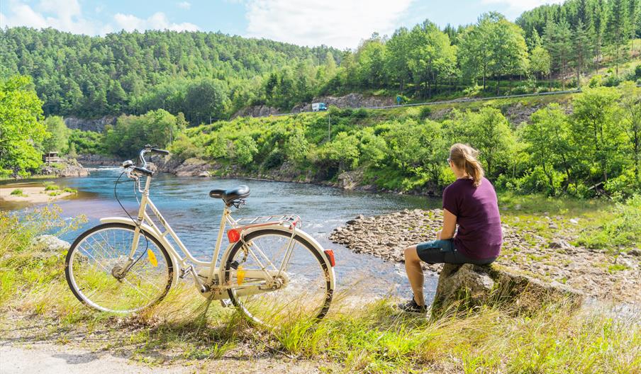 En dame sittende på en stein og ser mot Mandalselva, hvit sykkel vedsiden av henne.