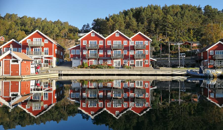 Tregde ferie, bilde tatt fra vannet inn mot brygga, røde bygninger med hvite karmer.