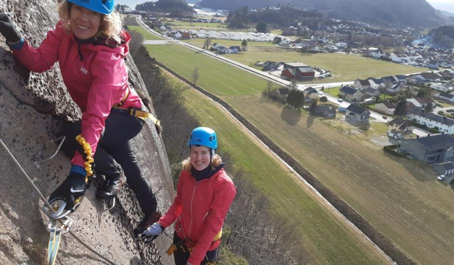To personer, iført sikkerhetsutstyr, klatrer Via Ferrata i Lyngdal. I bakgrunnen kan man se boligområdet, grønt landskap, og fjell.