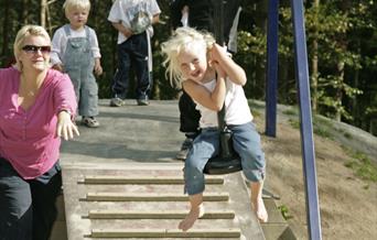 4 barn venter på tur til å ta en slynge, ei jente sitter på slyngen og ble dyttet av en voksen