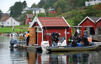 Bilde av en rød bod på en brygge med minst 8 dykkere som gjør seg klare for å komme seg på en svart båt på Tregde