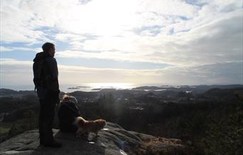 En mann som står og en dame som sitter ved en liten hund (tibetansk spaniel) ser utover mot husene, fjellet og havet. Sol bak skyene på himmelen