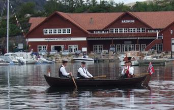Liten trebåt på vannet med 2 menn og 2 kvinner i bunader, og det norske flagget på tuppen av båten foran den røde Lindesnes Hytteservice bygningen.