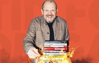 Komiker Freddy Kjensmo smilende med en stabel bøker som står i flammer foran seg.