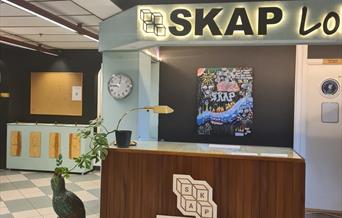 Resepsjonsdisk hos SKAP. Brun disk med SKAP logo på  Blågrå og hvite rutete gulv, blå og svart vegg med svære bokstaver på veggen "SKAP"