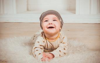Bilde av en baby liggende på magen, på et teppe på gulvet. Babyen kikker oppover og smiler. Foto: Vika_Glitter