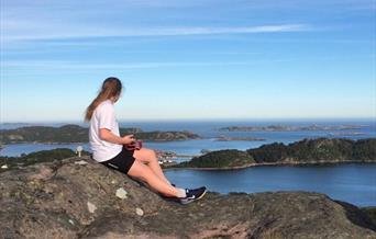 Bilde av en kvinne med hestehale som sitter på fjellet og ser ut mot havet, på havet er det flere fjell