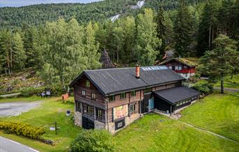 flyfoto av Hillestad galleri, en tømmerbygning omgitt av grønt gress og skog.