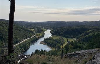 Bilde av landskap med elv midt i, vei langs vannet og skog på høyre side, få skyer på himmelen og fint vær