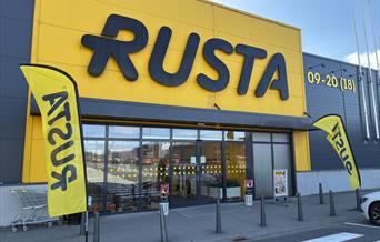 Inngangen til Rusta Lyngdal i Handelsparken, med gul fasade og flagg med logo.