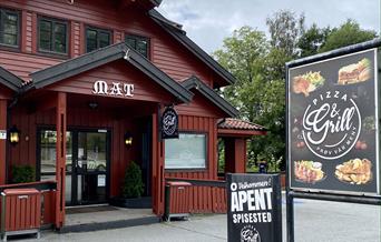 Delvis fasade av inngangen. Rød bygning med hvit skrift "mat" over inngangen. To store plakater med reklame av Mones Pizza & Grill.