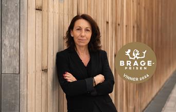 forfatter Kristina Quintano kledd i svart med armene i kors lener seg mot en trevegg. På bilde er også logo av Brageprisen vnner 2024.