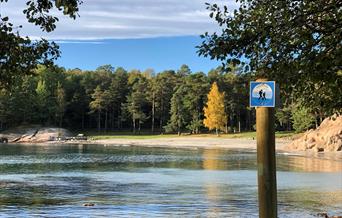 Bukt med strand og trær i bakgrunnen. Et liste skilt med "kyststien" fremme i bildet.