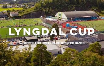 Oversiktsbilde av Lyngdal Stadion fylt med fotballbaner og mennesker under Lyngdal Cup. Midt i bildet står teksten "Lyngdal Cup – Minner for livet, på og utenfor banen". I bakgrunnen sees hall, tribuner og grønne omgivelser.